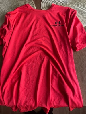 Orange Under Armour t-shirt - Snygg röd t-shirt från Under Armour med logga och text på bröstet. Klassisk rund halsringning och korta ärmar. Perfekt för dig som gillar sportig stil och vill ha något bekvämt och lättburet.