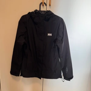 Svart vindjacka Helly Hansen XS - Svart vindjacka från Helly Hansen i storlek XS. Jackan har huva, dragkedja framtill och två fickor. Tillverkad i lätt och slitstark polyester, perfekt för blåsiga dagar. Klassisk HH-logga på bröstet och justerbar nederkant.
