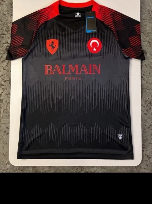 L Turkisk svart och röd fotbollströja  - Med pris lapp Bara köpte det va stor på mig har M trodde den skulle passa den är L Snygg svart och grå  med röda detaljer och balmain tryck. Tröjan har korta ärmar, grafiskt mönster, och loggor på bröstet som turkisk flagga och Ferrari På ryggen står det ASLAN, vilket betyder lejon på turkiska mit eget köpt design är hella tröjan nummer 7 och FLAWLESS i rött. Perfekt för träning eller match.