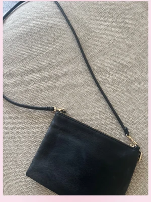 Svart axelväska i skinnimitation - 💕Snygg svart axelväska med enkel, stilren design och justerbar axelrem. Väskan har guldfärgade detaljer och dragkedja upptill. Perfekt för att bära det viktigaste och passar till de flesta outfits.💓