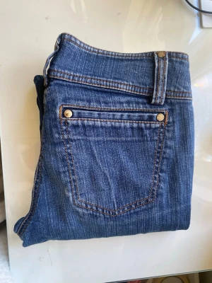 Blå bootcut jeans med dubbla knappar - Snygga blå bootcut jeans med dubbla knappar i midjan och guldfärgade nitar. Klassisk femficksmodell med kontrastsömmar och lätt utsvängda ben. Jeansen har en tidlös tvätt och är tillverkade i denim med bomullskänsla.