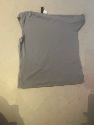 Ljusgrå axelbandslös tubtopp - Säljer en enkel och stilren ljusgrå tubtopp med bandeau-modell. Toppen är ärmlös och har en lätt rynkad detalj upptill för bättre passform. Perfekt att matcha med jeans eller kjol för en clean look. Andrig Andvänt men pris lappen är borta 