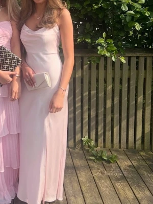 Ljusrosa balklänning  - Elegant ljusrosa satin långklänning och lätt draperad urringning. Klänningen har en diskret slits framtill och figurnära passform som framhäver siluetten. Perfekt för bal. Använd 1 gång 