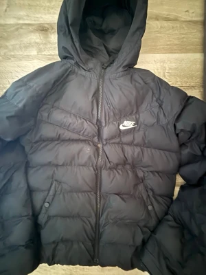 Svart pufferjacka från Nike - Säljer en svart pufferjacka från Nike med huva och dragkedja framtill. Jackan har två fickor med tryckknappar och ett broderat Nike-logotyp på bröstet. Perfekt för kalla vinterdagar och riktigt skön att bära. Materialet är slitstarkt och håller värmen bra.