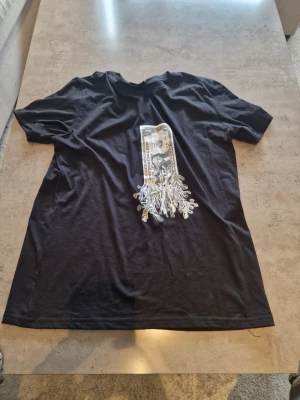 Svart t-shirt med dollartryck - Svart t-shirt med ett unikt tryck av en dollarsedel som smälter ner över bröstet. T-shirten har rund hals och korta ärmar, perfekt för en streetwear-look. Materialet känns mjukt och bekvämt mot huden. Storlek small men passar stort