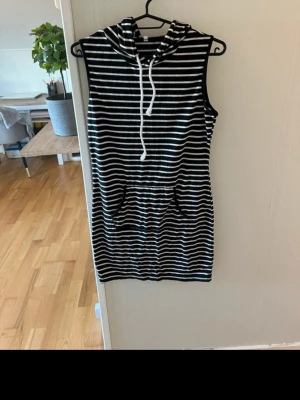 Randig ärmlös hoodieklänning - Säljer en svartvit, ärmlös hoodieklänning med huva och snörning. Klänningen har ett randigt mönster och en ficka framtill. Perfekt för dig som gillar en avslappnad och sportig stil.