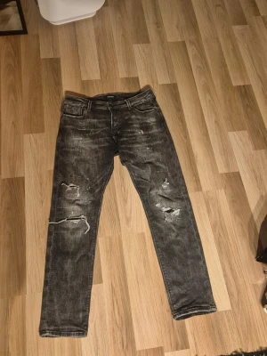Grå slitna jeans med hål - Säljer ett par grå jeans med slitningar och hål på båda benen. Jeansen har fem fickor, raka ben och en cool, tvättad look. Perfekta för dig som gillar en avslappnad och trendig stil.