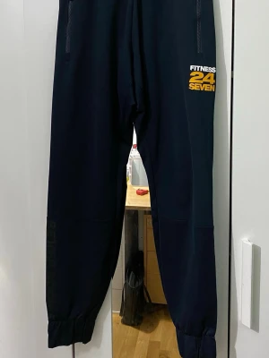 Svarta Better Bodies mjukisbyxor - Mörkblå mjukisbyxor från Better Bodies med tryck i gult och vitt från Fitness 24 Seven på ena benet. Byxorna har resår i bensluten och dragkedjefickor. Perfekta för gymmet eller chill hemma. Mått: längden är 96cm och midjan är 34cm. 