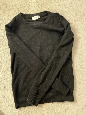 Svart stickad tröja från Vila - En enkel och stilren svart stickad tröja från Vila. Tröjan har rund halsringning, långa ärmar och ribbade muddar vid ärmslut och nederkant. Perfekt basplagg att matcha med olika outfits.