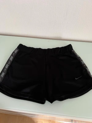 Svarta Nike shorts med logga - Snygga svarta shorts från Nike med elastisk midja och logga längs sidorna. Tillverkade i ett glansigt syntetmaterial som känns sportigt och bekvämt. Perfekta för träning eller chill dagar.