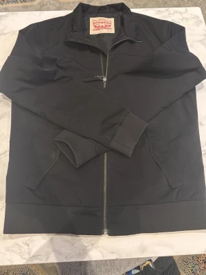 Svart vindjacka från Levi's - Snygg svart vindjacka från Levi's med dragkedja framtill och ståkrage. Jackan har ribbade muddar vid ärmslut och nederkant samt två sidofickor. Tillverkad i ett lätt och slitstarkt material som passar perfekt till streetwear-stilen, pass form är till Man