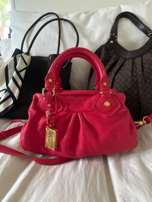 Röd Marc by Marc Jacobs handväska - Så cool rosaröd handväska i äkta läder från Marc by Marc Jacobs. Den ikoniska modellen Q Classic Baby Groove! En populär och eftertraktad vintagemodell med gulddetaljer och dubbla handtag. Långa bandet finns. Så fint skick och en otroligt snygg vintage väska🤍  Mått: 34x18x12 cm (undersida) Kan hämtas hemma hos mig i Nacka eller skickas 🤍