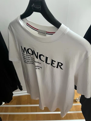Vit Moncler t-shirt med tryck - Snygg vit t-shirt från Moncler med stort svart tryck på bröstet och textdetaljer. Klassisk rund halsringning och korta ärmar. Tillverkad i mjuk bomull som känns skön mot huden. Perfekt för dig som gillar stilrena plagg med exklusiv känsla.