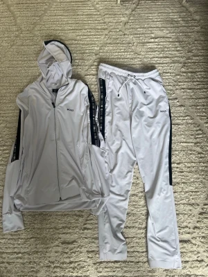 Vit loungewear-set från BOSS - Snyggt vitt loungewear-set från BOSS med matchande hoodie och byxor. Setet har svarta detaljer längs ärmar och ben med BOSS-logga, dragkedja på hoodien och dragsko i midjan på byxorna. Tillverkat i mjukt och stretchigt material för maximal komfort. Storlek:Small. Passar om du är runt 175.