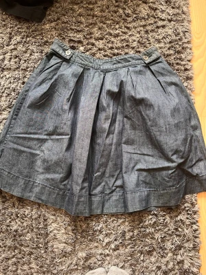 Mörkblå denimkjol med veck - Säljer en mörkblå kjol i denim med snygga veck framtill och dekorativa knappar vid linningen. Kjolen har en lätt utställd form och är gjord i ett kraftigare bomullstyg som ger en cool och avslappnad look.