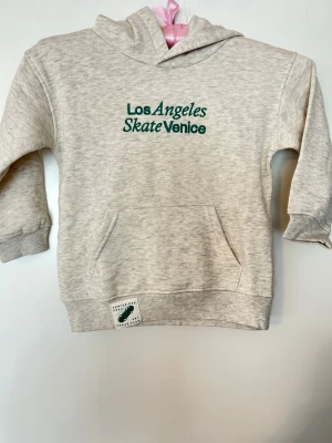 Grå hoodie Los Angeles Skate Venice - Ljusgrå hoodie med grön brodyrtext 'Los Angeles Skate Venice' på bröstet. Klassisk känguruficka framtill och ribbade muddar vid ärmar och nederkant. Mjuk och skön, perfekt för chill dagar. Liten patch nertill på magen. Materialet känns som bomullsblandning.