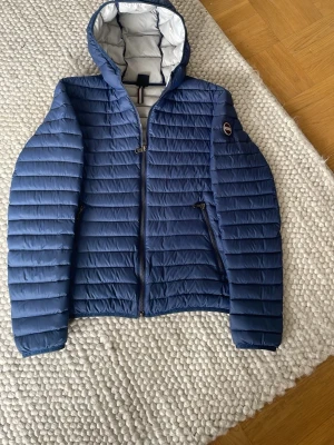 Blå pufferjacka med huva från Colmar - Snygg blå pufferjacka från Colmar med huva och vita detaljer på insidan. Jackan har två dragkedjeförsedda fickor och Colmar-logga på ärmen. Lättviktigt material och quiltad design gör den både stilren och praktisk för kalla dagar. Nypris cirka 3200kr , den är knappt använd och är väldigt fin och perfekt nu till våren / sommaren och även hösten! Hör av er vid minsta fundering 