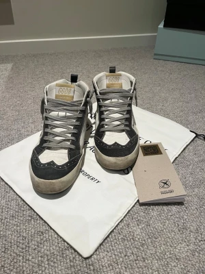 Golden Goose Mid Star sneakers grå/vit - Golden Goose Mid Star sneakers i grått och vitt med ikonisk stjärndetalj på sidan. Skorna har grå mockadetaljer, vita partier i läder och grå snörning. Klassisk logga på plösen och svart hälflik. Snygg vintage-look och cool design.