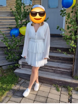 Studentklänning / sommarklänning  - Studentklänning / sommarklänning.             Säljer en superfin vit kortklänning med omlottdesign och volangdetaljer. Klänningen har långa, vida ärmar och v-ringning med knyt framtill. Perfekt för sommarens alla tillfällen och studentfirande! Endast använd en gång så i riktigt bra skick. Storlek 36.    Kolla gärna in resten av min garderob där det bland annat finns flera klänningar, jeans mm