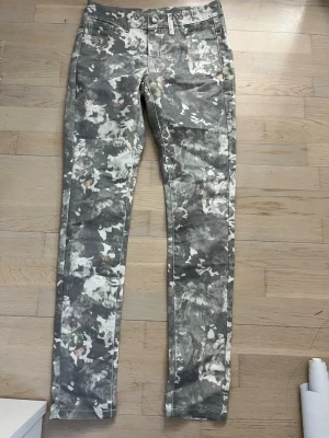Gråblommiga jeans med raka ben - Unika jeans med grå och vita blommönster samt inslag av beige och grönt. Skinnet jeans och klassisk femficksmodell. Snyggt tryck som sticker ut och passar perfekt till en trendig streetstyle-look. Materialet känns som bomull med stretch.