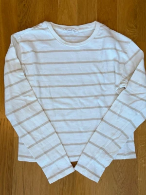 Randig långärmad topp från Mango - Snygg långärmad topp från mango i vitt med beigea horisontella ränder. Klassisk rund halsringning och mjukt material som känns skönt mot huden. Perfekt att styla med jeans eller kjol för en enkel och fräsch look.
