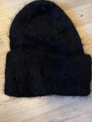 Svart fluffig stickad mössa - Säljer en svart, fluffig stickad mössa med uppvikt kant. Mössan har en enkel och klassisk form och är tillverkad i ett mjukt, lurvigt material som ger en mysig känsla. Perfekt för kalla dagar och passar till de flesta stilar.
