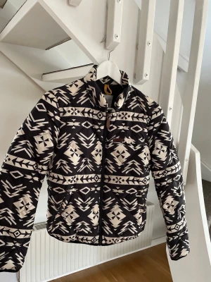 Svartvit mönstrad pufferjacka - Säljer en svartvit pufferjacka med grafiskt mönster och hög krage. Jackan har dragkedja framtill och två sidofickor. Perfekt för kalla dagar och ger en riktigt snygg streetwear-look. Materialet är vadderat och håller värmen bra.