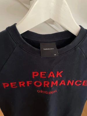 Mörkblå sweatshirt Peak Performance - Mörkblå sweatshirt från Peak Performance med röd broderad logga och texten 'ORIGINAL' framtill. Klassisk rund halsringning och ribbade muddar vid ärmslut och nederkant. Perfekt för en avslappnad och sportig stil.