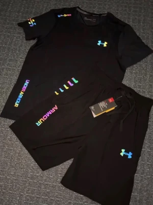 Svart träningsset från Under Armour - Snyggt svart träningsset från Under Armour med t-shirt och shorts. Setet har färgglada loggor och detaljer i regnbågsfärger på både tröja och byxa. Perfekt för gymmet eller löprundan. Materialet är lätt och andas, vilket gör det bekvämt under träning.