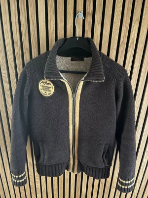 Mörkbrun stickad kofta med dragkedja - Mörkbrun stickad kofta med grov ribbad krage och dragkedja framtill. Beige detaljer längs dragkedjan och på ärmsluten. Patch med text och bergmotiv på bröstet. Två fickor fram och breda muddar. Perfekt för dig som gillar retrovibbar och mysig stil.