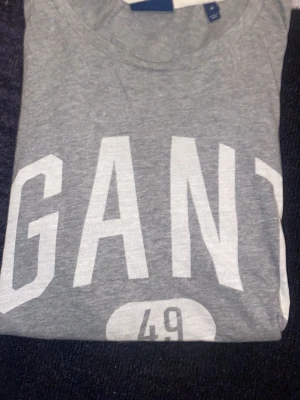 Grå GANT t-shirt med tryck - Snygg grå t-shirt från GANT med stort vitt tryck framtill där det står 'GANT 49 NEW HAVEN'. Klassisk rund halsringning och mjukt material, perfekt för en avslappnad stil. Passar dig som gillar sportig och enkel design.