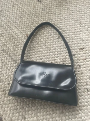 Vintage väska❤️ - Snygg svart vibtage axelremsväska  i glansigt läder. Klassisk rektangulär form med lock och diskret logga på framsidan. Perfekt storlek för det viktigaste och enkel att bära över axeln. En tidlös accessoar som lyfter vilken outfit som helst.