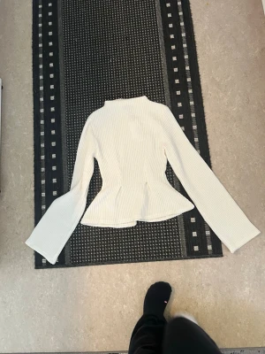 Vit ribbad topp med trumpetärm - Trendig vit ribbad topp med lång trumpetärm och hög hals. Figurnära passform med markerad midja och lätt utsvängd nederkant. 