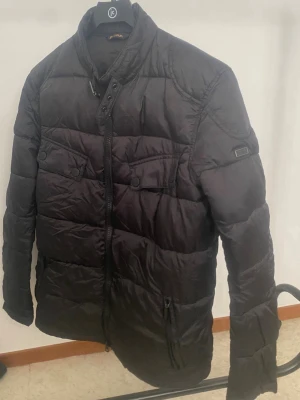 Barbour International svart jacka, storlek S - Snygg svart dunjacka med hög krage och tryckknappar. Jackan har två bröstfickor med lock och knappar samt dragkedjefickor framtill. Quiltad design och diskret logga på ärmen. Perfekt för kalla vinterdagar.