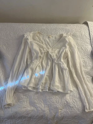 Vit blus med volang och v-ringning - Romantisk vit blus med v-ringning och volangdetaljer längs bröstet och axlarna. Långärmad modell i tunt, lätt material som ger en luftig känsla. Perfekt för dig som gillar en feminin och trendig stil.