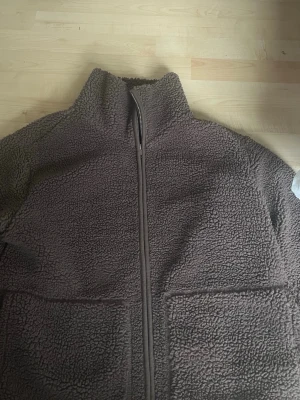 Brun teddyjacka med dragkedja - Mysig brun teddyjacka med hög krage och hel dragkedja framtill. Jackan har två stora fickor framtill och är gjord i fluffigt teddy-material som håller dig varm. Perfekt för kyliga dagar och ger en avslappnad look.