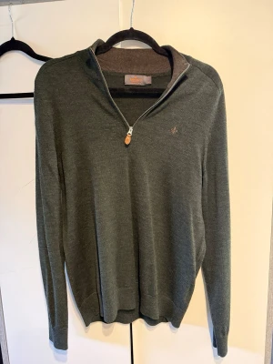 Mörkgrön halvzip-tröja från Morris  - Mörkgrön stickad tröja från Morris i Marino Wool, med halvzip och liten broderad logga på bröstet. Tröjan har ribbade muddar vid ärmslut och nederkant samt en högre krage med dragkedja och läderdetalj på zippen. Perfekt för en stilren look.