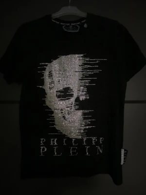 Svart t-shirt med döskalle från Philipp Plein - Svart t-shirt från Philipp Plein med glittrande döskallemotiv framtill och märkets logga i silver. Klassisk rund halsringning och korta ärmar. Materialet känns mjukt och skönt, troligen bomull. Perfekt för dig som gillar edgy och unik stil. Äkta