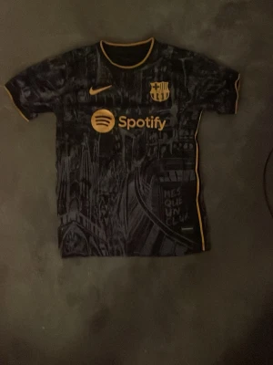 Barcelona matchtröja svart/guld Nike - Snygg svart Barcelona matchtröja med guldiga detaljer och tryck från Spotify och UNHCR. Tröjan har korta ärmar, rund hals och coolt grafiskt mönster inspirerat av staden. Perfekt för dig som älskar fotboll och vill sticka ut på planen. Väldigt bra skick då den har knappt varit använd 