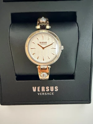 Guldig klocka från Versus Versace - Stilren klocka från Versus Versace i guld med vit urtavla och elegant mönster. Armbandet har dekorativa detaljer med gnistrande stenar. Perfekt accessoar för dig som vill ha något lyxigt och trendigt på handleden.