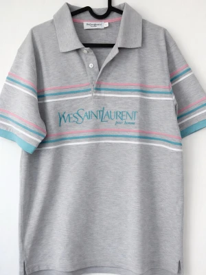 Grå randig piké från Yves Saint Laurent - Säljer en grå piké från Yves Saint Laurent med turkosa och rosa ränder samt tryckt logga på bröstet. Skjortan har korta ärmar, klassisk krage och knappar vid halsen. Perfekt för dig som gillar retrovibbar och stilren design.
