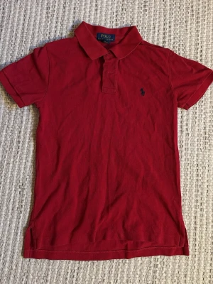Röd pikétröja från Polo Ralph Lauren - Storlek barn 11-12. 146-152. Klassisk röd pikétröja från Polo Ralph Lauren med krage, korta ärmar och den ikoniska broderade loggan på bröstet. Tröjan har en slim passform och är tillverkad i mjuk bomull som känns skön mot huden.