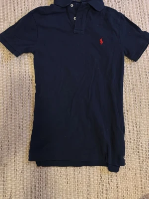 Marinblå pikétröja från Polo Ralph Lauren - Klassisk marinblå pikétröja från Polo Ralph Lauren med röd broderad logga på bröstet. Tröjan har korta ärmar, krage och knappar vid halsen. Tillverkad i mjuk bomull och har en slim fit passform.