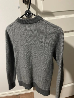 Grå stickad tröja med fiskbensmönster - Snygg grå stickad tröja med fiskbensmönster och mörkare ribbade muddar vid hals, ärmslut och nederkant. Tröjan har rund halsringning och lång ärm, perfekt för dig som gillar stilrena och klassiska plagg.