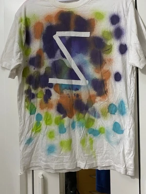 Vit t-shirt med färgglad spraydesign - Unik vit t-shirt med handgjord sprayad design i lila, orange, grön och blå toner. Stor vit symbol mitt på bröstet ger en cool och kreativ vibe. T-shirten har korta ärmar och avslappnad passform, perfekt för dig som gillar streetwear och konstnärliga plagg.