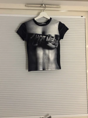 Svart croppad t-shirt med tryck - Snygg svart croppad t-shirt med ett grafiskt tryck framtill där det står 'HOT MESS'. T-shirten har korta ärmar och är figurnära i modellen. Materialet känns som polyester och ger en lite glansig finish. Perfekt för dig som vill sticka ut!