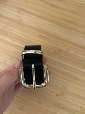 Svart bälte med silverspänne - Stilrent svart bälte med ett rejält silverspänne. Bältet har en klassisk design och är tillverkat i slätt skinn. Perfekt för att lyfta vilken outfit som helst med en enkel men snygg detalj.