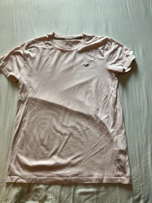 Ljusrosa t-shirt från Hollister - Säljer en ljusrosa t-shirt från Hollister med klassisk rund hals och diskret logga på bröstet. T-shirten är kortärmad och gjord i mjuk bomull, perfekt för en avslappnad och stilren look.