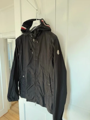 Svart Moncler Windbreaker - Snygg svart vindjacka från Moncler med huva och dragkedja framtill. Jackan har Moncler-logga på ärmen och detaljer i rött, vitt och svart på huvan. Tillverkad i lätt och tåligt material, perfekt för blåsiga dagar. Stolek 3 = M/L.  Äkta.