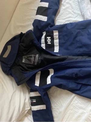 Blå Helly Hansen vindjacka med huva - Snygg marinblå vindjacka från Helly Hansen med vita och svarta detaljer på ärmar och bröst. Jackan har huva, dragkedja och HH-logga framtill. Perfekt för blåsiga dagar och sportiga outfits.